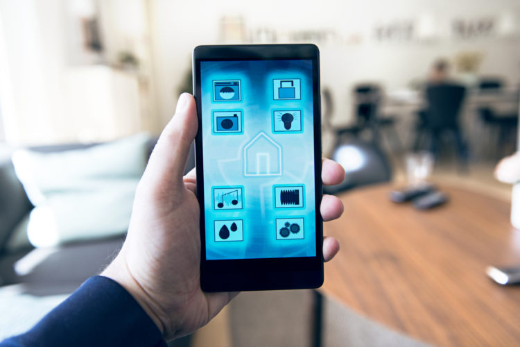 Point of view of hand that holds a mobile phone with an app to control wireless devices in a smart home like washing machine, security camera, door lock, music player and much more. Concept of IOT (Internet of Things).