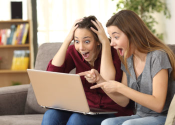 Two astonished roommates watching media content on line with a laptop sitting on a couch in the living room in a house interior