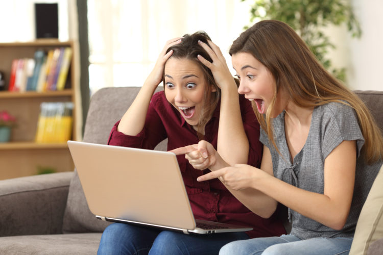 Two astonished roommates watching media content on line with a laptop sitting on a couch in the living room in a house interior